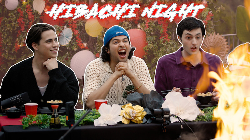 From left to right, Tanner Buchanan, Xolo Maridueña, and Gianni DeCenzo are seated at a dinner table, with flames rising dramatically on the right side.