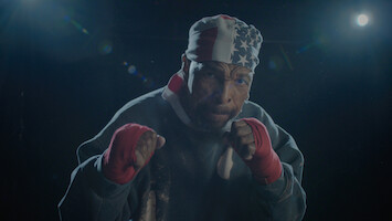 A male boxer wearing red hand wraps and a USA flag bandana poses in a fighting stance under dramatic lighting in a dark gym or training environment.