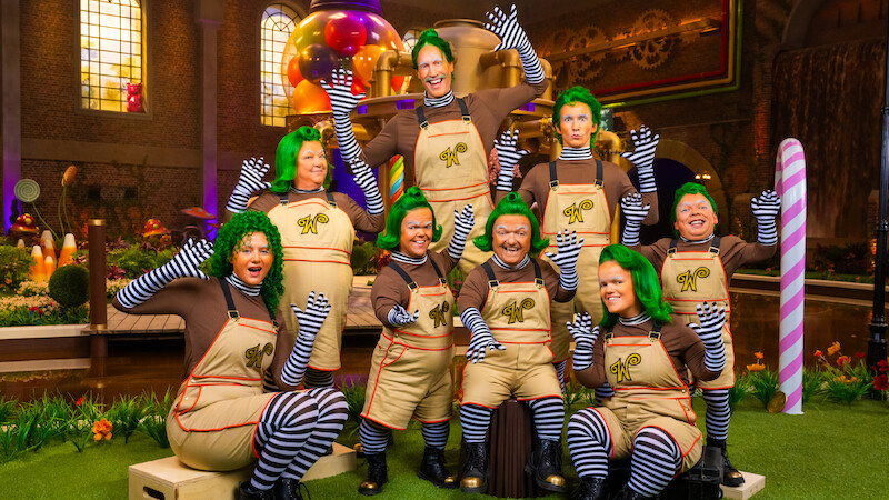 Grupo de pessoas fantasiadas de umpa-lumpas, com perucas verdes, camisas marrons, leggings listradas e macacões, no cenário de uma fábrica de doces colorida e extravagante, com guloseimas gigantes e iluminação vibrante.