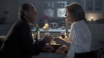 A man and woman sit at a kitchen table having dinner with wine, surrounded by lit candles, sharing a warm, intimate moment in a softly lit home environment.