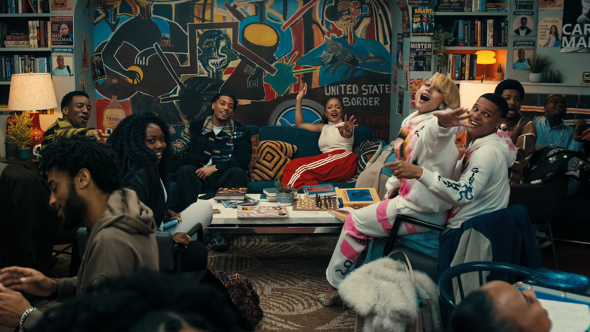Um grupo diverso de pessoas ri e conversa em uma sala de estar colorida repleta de obras de arte, livros e um grande mural, criando uma atmosfera vivaz e receptiva.