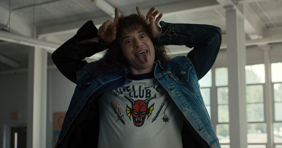 Eddie Munson (Joseph Quinn) stands indoors, playfully sticking out his tongue and making horn gestures with his hands while wearing a "Hellfire Club" t-shirt and denim jacket.