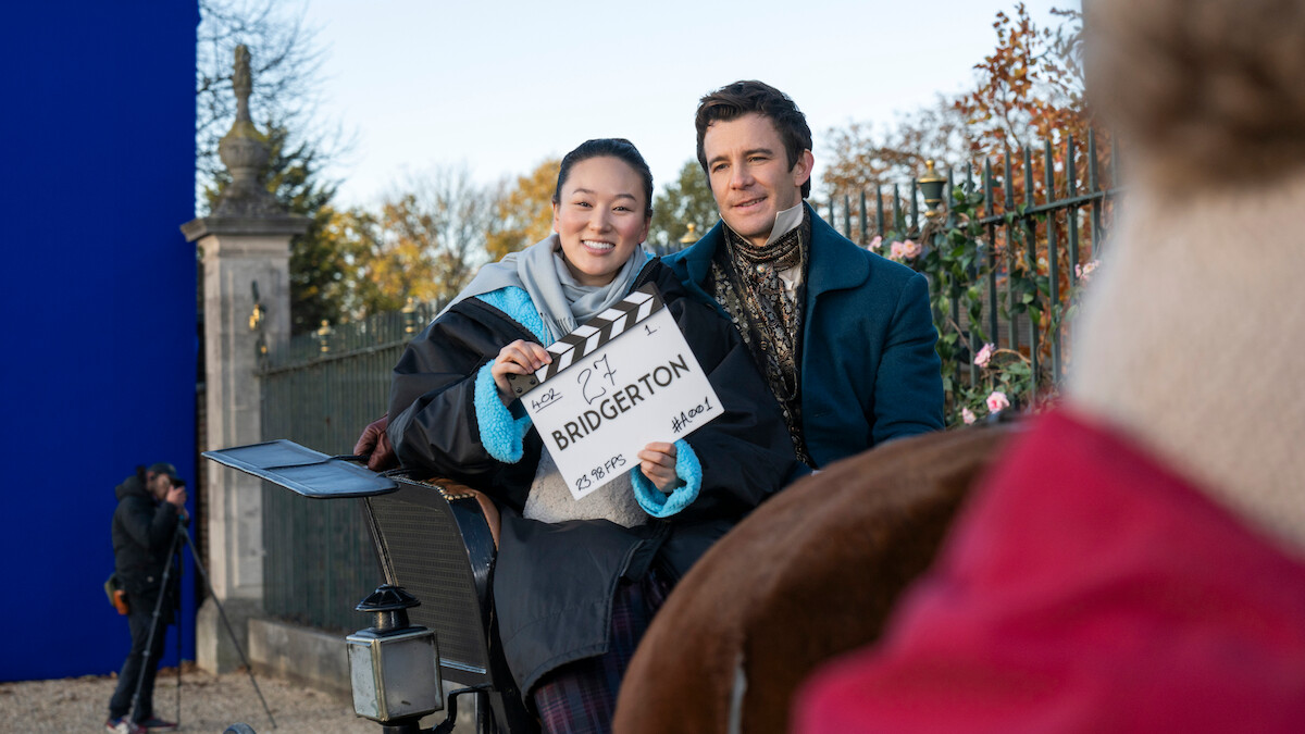 Two people in period costumes sit in a carriage on an outdoor film set, holding a "Bridgerton" clapperboard, with a blue screen and autumn trees in the background.