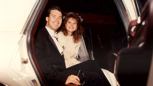 A smiling bride and groom in formal wedding attire sit together in the back seat of a car, looking joyful and relaxed, suggesting a celebratory and happy atmosphere.