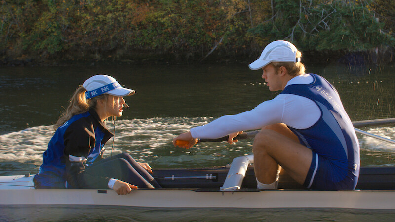 Two rowers in blue uniforms and white caps sit in a racing shell on a calm river, surrounded by greenery, appearing to be in discussion or preparing for a rowing session.