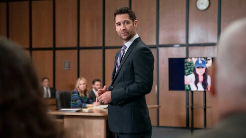 Man in suit stands in courtroom, addressing others. People sit at a table in the background. A digital screen shows a woman’s photo. Wood panel walls and a clock indicate a formal court setting.