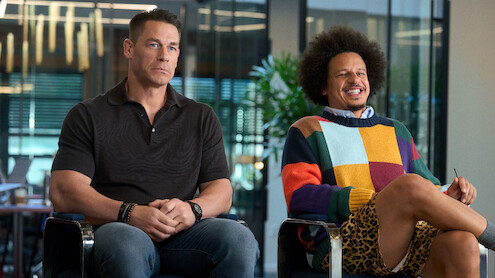Two men sit indoors on chairs, one in a black polo shirt looking serious, the other in a colorful sweater and animal-print shorts smiling, with plants and modern office glass walls in the background; mood is contrasting and casual.