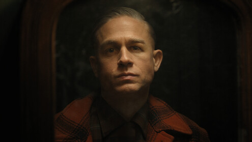 Ed Gein in a plaid jacket looks at his reflection in the mirror in a dark room.