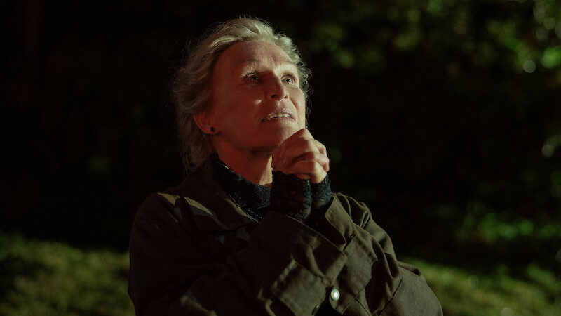 An older woman stands outdoors at night, hands clasped in front of her face as if in prayer, looking upward with a hopeful and contemplative expression, illuminated by warm light against a dark, blurred background.