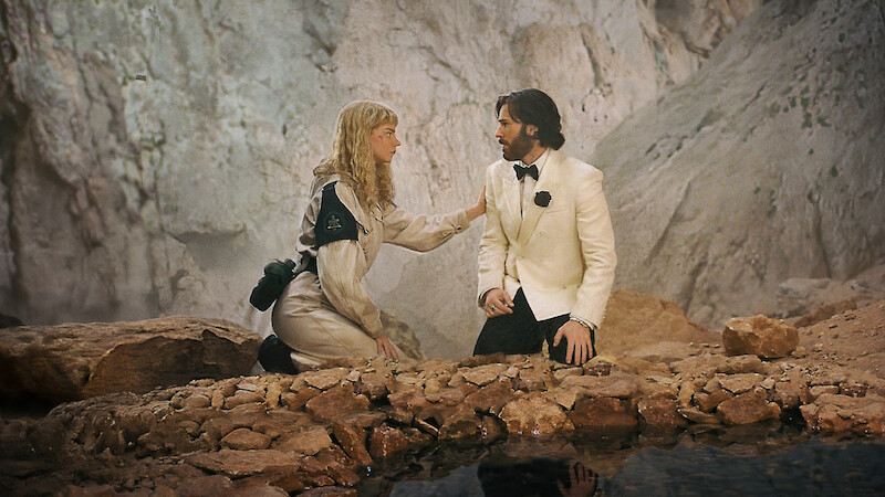 A woman in an adventure outfit kneels by a man in a white tuxedo; they appear to be talking in a rocky, cave-like environment near a shallow pool of water.