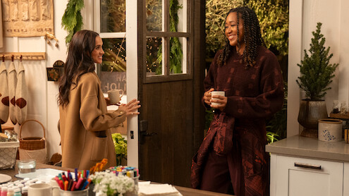 Two women smiling and talking in a cozy kitchen decorated for the holidays, holding mugs. Warm lighting, craft supplies on the counter, and greenery create a welcoming, festive atmosphere.