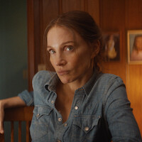 Woman with light brown hair in a denim shirt sits indoors, leaning on a wooden chair. Wooden paneled wall and framed photos are visible in the warm-lit background, creating a cozy home setting.