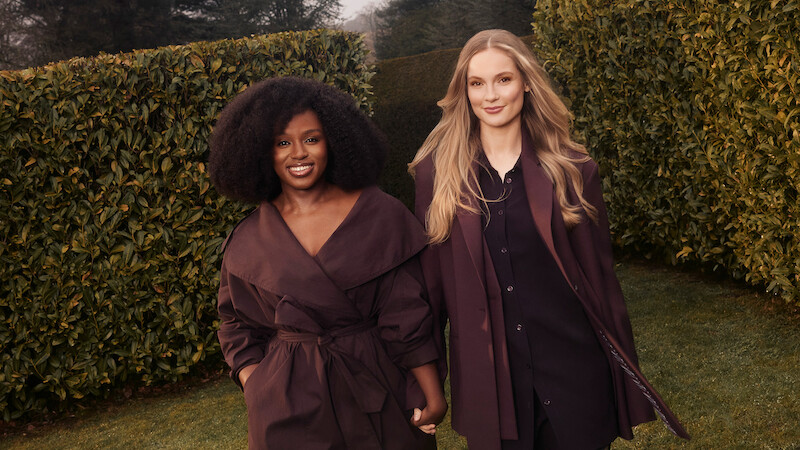Two women standing outdoors in a garden with tall manicured hedges, wearing stylish, coordinated dark outfits, smiling and holding hands, with soft natural lighting and trees in the background.