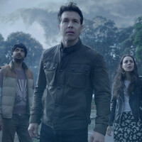Three people stand outdoors, looking up with concerned expressions. Behind them are tall trees and a cloudy, stormy sky, suggesting an intense or dramatic situation in a natural environment.