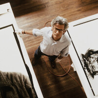A man with glasses stands on a wooden floor between large white panels, looking up at the camera. Artworks and a coiled cable are visible, suggesting a studio or creative workspace environment.