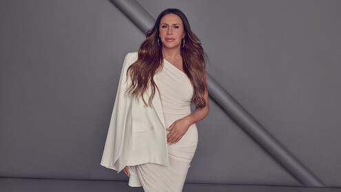 Karla Sofía Gascón wears a white one-shouldered dress against a grey background.