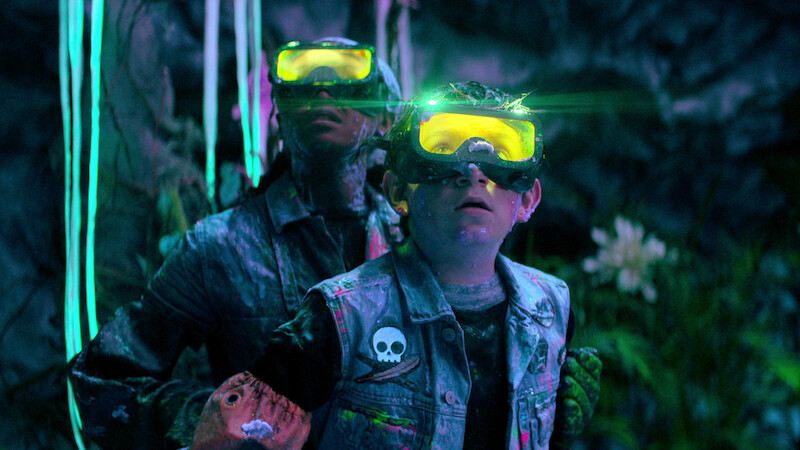 Two people wearing yellow glowing goggles and denim jackets with patches stand in a colorful, neon-lit cave environment, surrounded by plants and glowing decorations, looking upward as if intrigued or cautious.