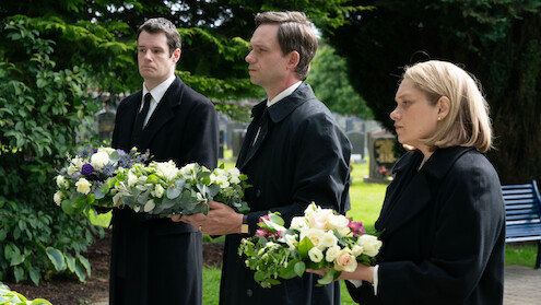 Three people in black funeral attire hold flower arrangements at a cemetery, standing solemnly among greenery and gravestones, with a blue bench in the background.