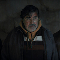 An older man with glasses and a beard, wearing a hoodie and jacket, stands indoors against a dimly lit, textured wall, looking slightly to the side.