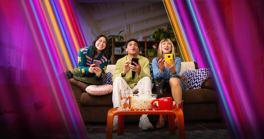 Three friends sit on a couch in a cozy living room, smiling and holding smartphones, with snacks on an orange table and colorful rainbow light effects creating a lively, cheerful atmosphere.