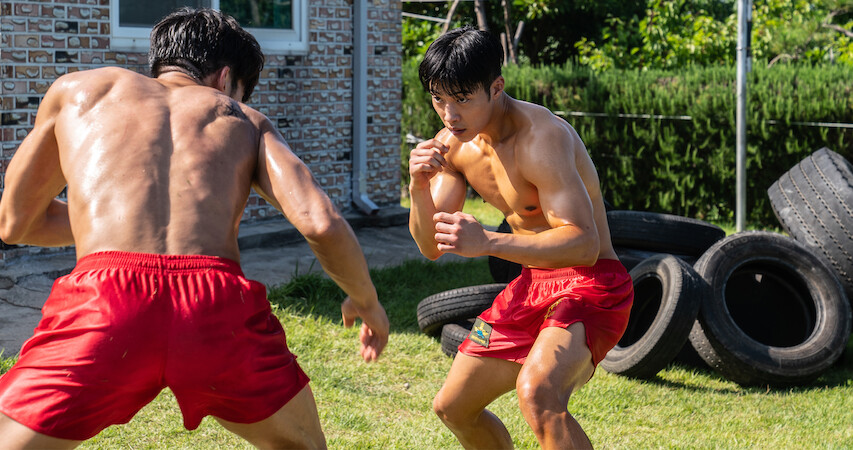 Two muscular  boxers in red shorts square off on a lawn with old tires stacked up behind them. 