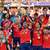 Soccer team in red jerseys celebrates on field, raising a gold trophy and wearing medals with confetti falling, showing excitement and unity after a championship victory.