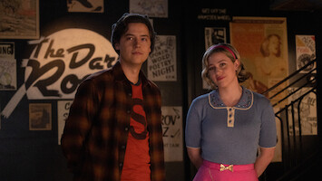 Cole Sprouse as Jughead Jones and Lili Reinhart as Betty Cooper stand together smiling in Season 7 of 'Riverdale.'