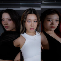 Three women pose indoors under low lighting, side by side, each with confident expressions; the background is dark and features horizontal metal slats.