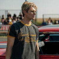 Young man with blond hair wearing a "Heavy Transport" T-shirt stands next to a red car at an outdoor event with spectators and a metal fence visible in the background.