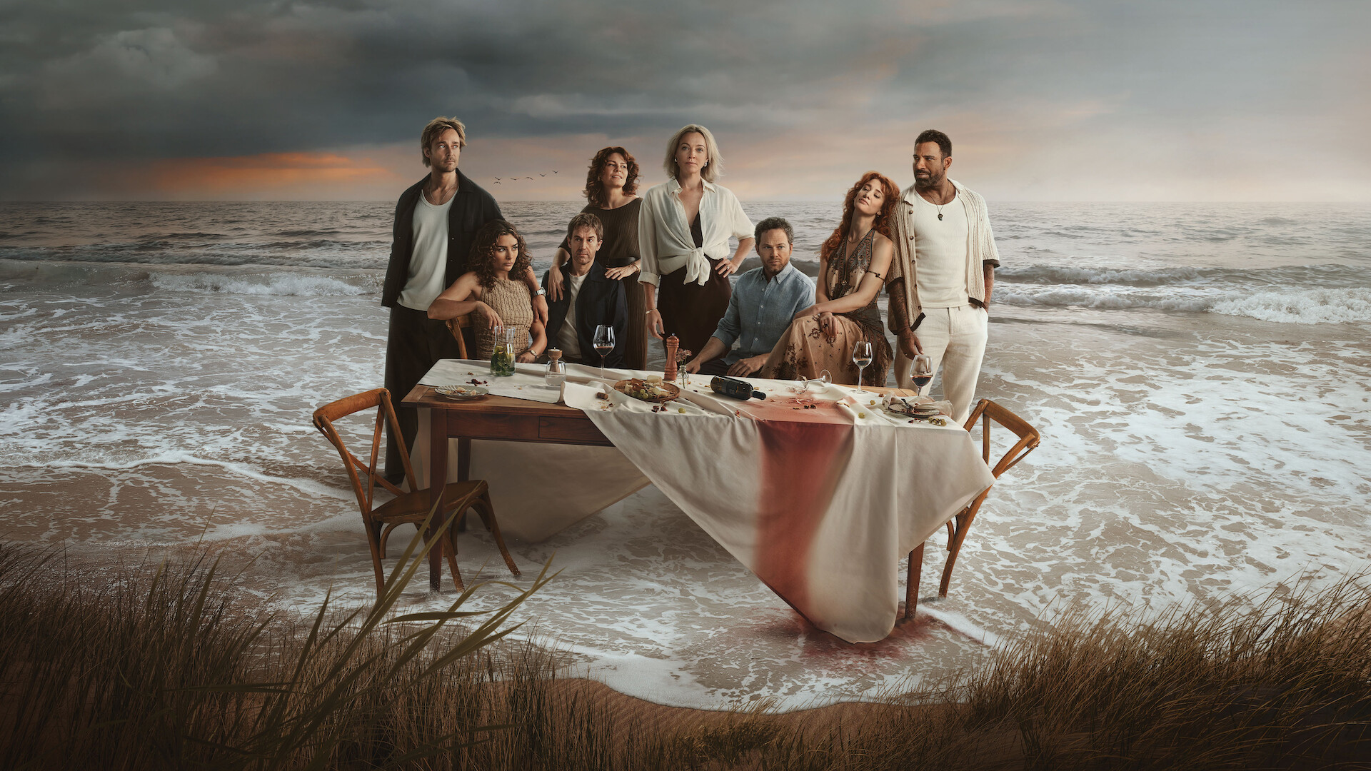 Eight people pose around a table with food and drinks on a beach at sunset, with dramatic waves and clouds behind them. The tablecloth is stained red, suggesting a mysterious or dramatic atmosphere.