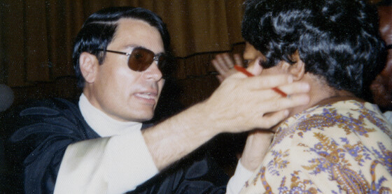 Man in glasses and dark robe extends his hands toward another person in a patterned shirt, indoors with brown curtain backdrop, suggesting an intense or dramatic interaction in a formal or ceremonial setting.
