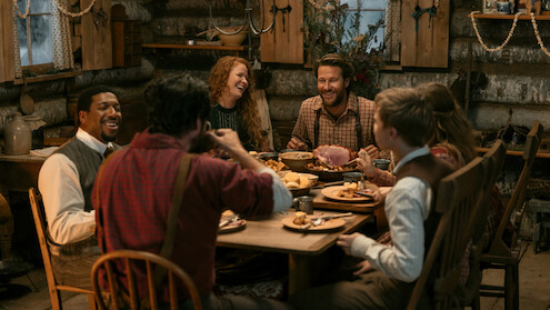 Six people in rustic clothing share a meal and laugh together inside a cozy log cabin, surrounded by wooden furniture, dim lighting, and a festive, warm atmosphere.