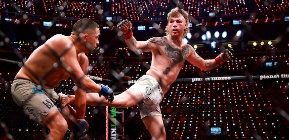 Two MMA fighters compete in a cage under bright arena lights; one delivers a high kick while the other defends, surrounded by a cheering audience and illuminated electronic displays.