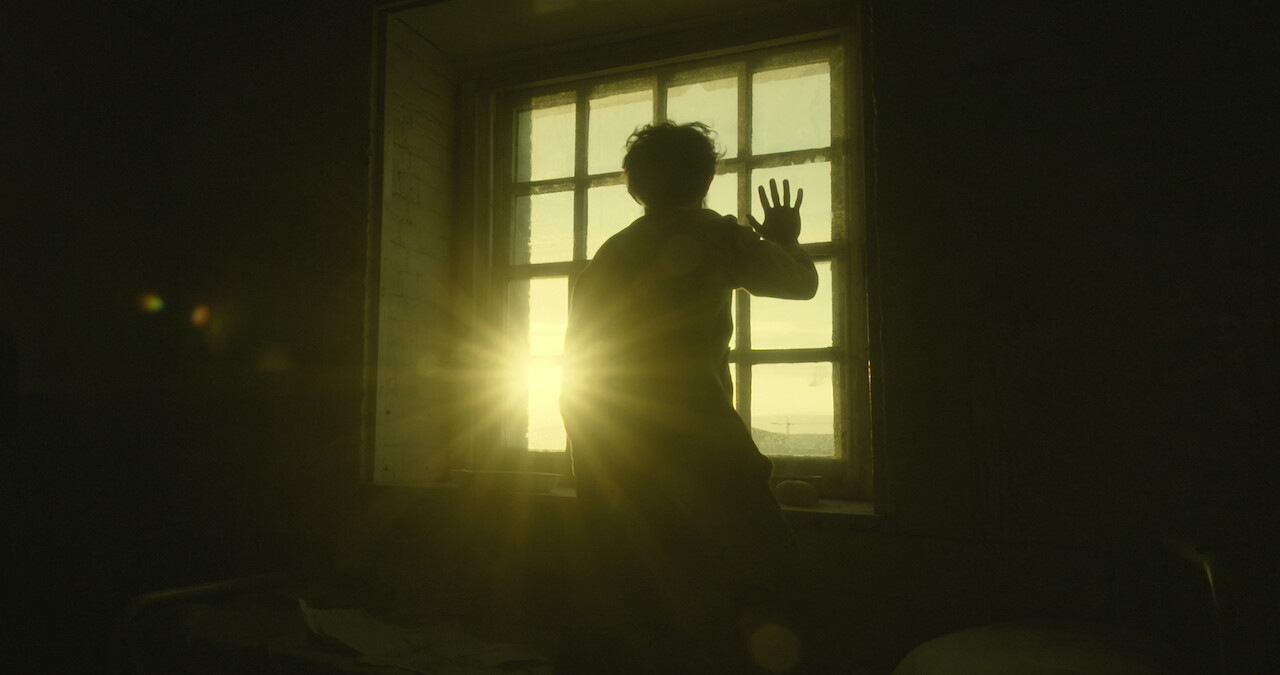   A man puts his hand to a window as the sun rises in the distance.
