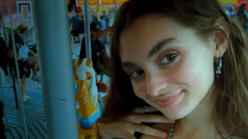 Young woman smiling for a selfie on a merry-go-round with carousel horse and people in the background, indoor amusement setting, colorful and lively atmosphere.