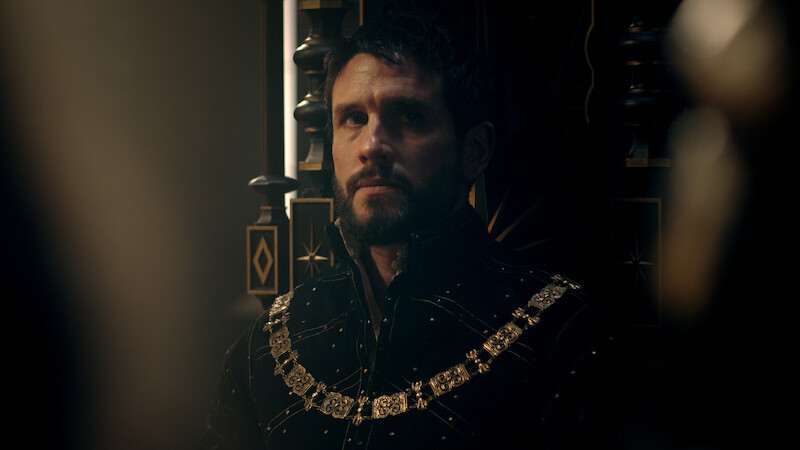 A solemn man in elaborate medieval attire with a gold chain sits on a dark, ornate throne in a dimly lit, regal setting, creating a moody, contemplative atmosphere.