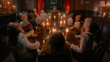 Jamie Campbell Bower como Henry Creel e um grupo de crianças ao redor de uma mesa iluminada por velas em um cômodo escuro em ‘Stranger Things 5’