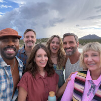 Six smiling adults taking a group selfie outdoors with mountains, desert, and cloudy sky in the background. They appear happy and relaxed, dressed in casual clothing, enjoying a scenic nature outing together.