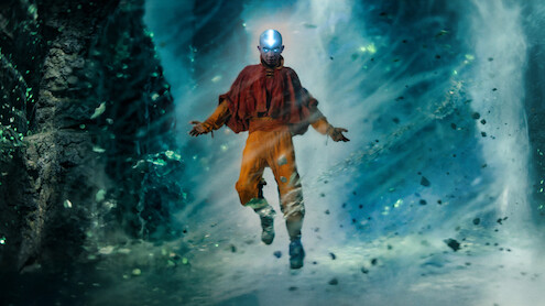 Gordon Cormier as Aang in traditional orange and red robes hovers in the air with glowing light on their head, surrounded by swirling wind and floating debris in a mystical cave or cavern environment.