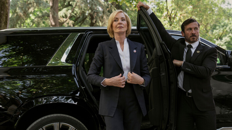 A woman in a suit stands confidently beside a black SUV with a security detail holding the door open, set outdoors with trees and greenery in the background, suggesting a formal or official environment.