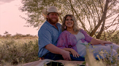 Smiling couple sitting on a blanket outdoors in a grassy field, with trees in the background, wearing casual clothes and cowboy hat, enjoying a relaxed moment in nature during daylight.