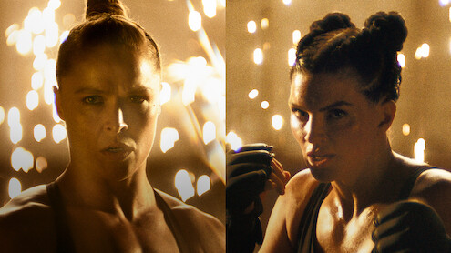 Ronda Rousey and Gina Carano with intense expressions face forward in a dramatic, dimly lit environment with sparks flying in the background, both wearing athletic gear and preparing to fight.