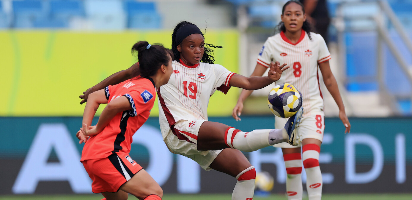 Female soccer players in action during a match, one in a white uniform controls the ball with her foot as opponents and teammates watch, in a stadium with blue seats and a colorful background.