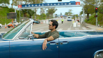 Man driving a classic blue convertible near Dodger Stadium entrance on a sunny day, with a relaxed vibe, palm trees and stadium signs visible, scene feels casual and laid-back.