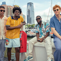 Four men in stylish summer outfits pose at a marina with boats, water, and tall city buildings in the background, suggesting a relaxed, urban waterfront setting.