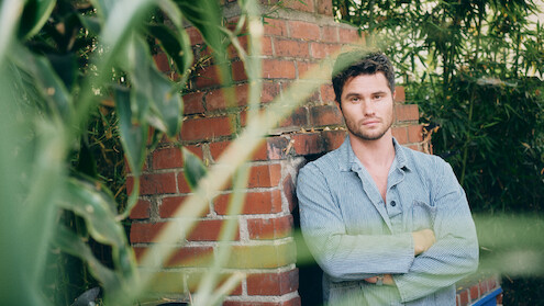 A man in a light denim shirt stands with arms crossed in front of a brick wall outdoors, surrounded by green plants and bamboo. The setting is natural and relaxed, with leaves partially obscuring the foreground.