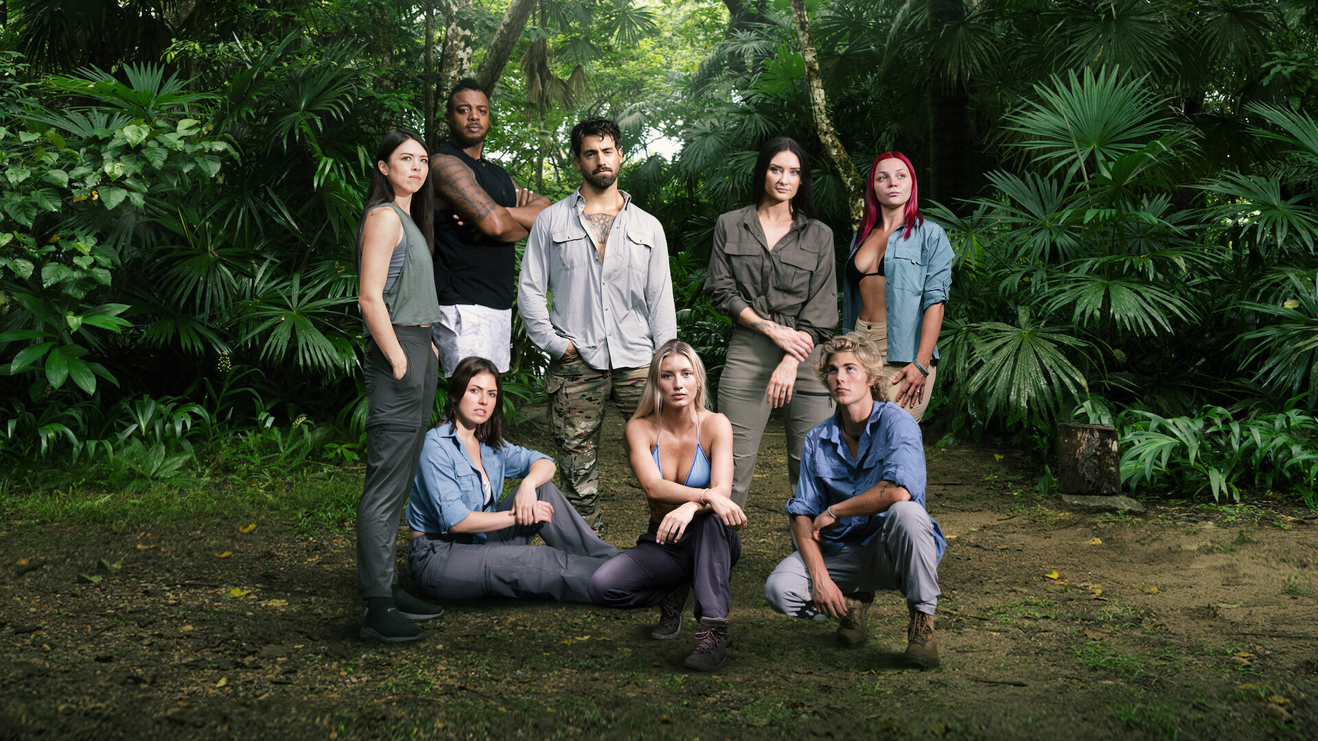 Group of eight people posing in a jungle setting, surrounded by lush green foliage, wearing outdoor adventure clothing, with some standing and others sitting on the ground, creating a team or expedition atmosphere.