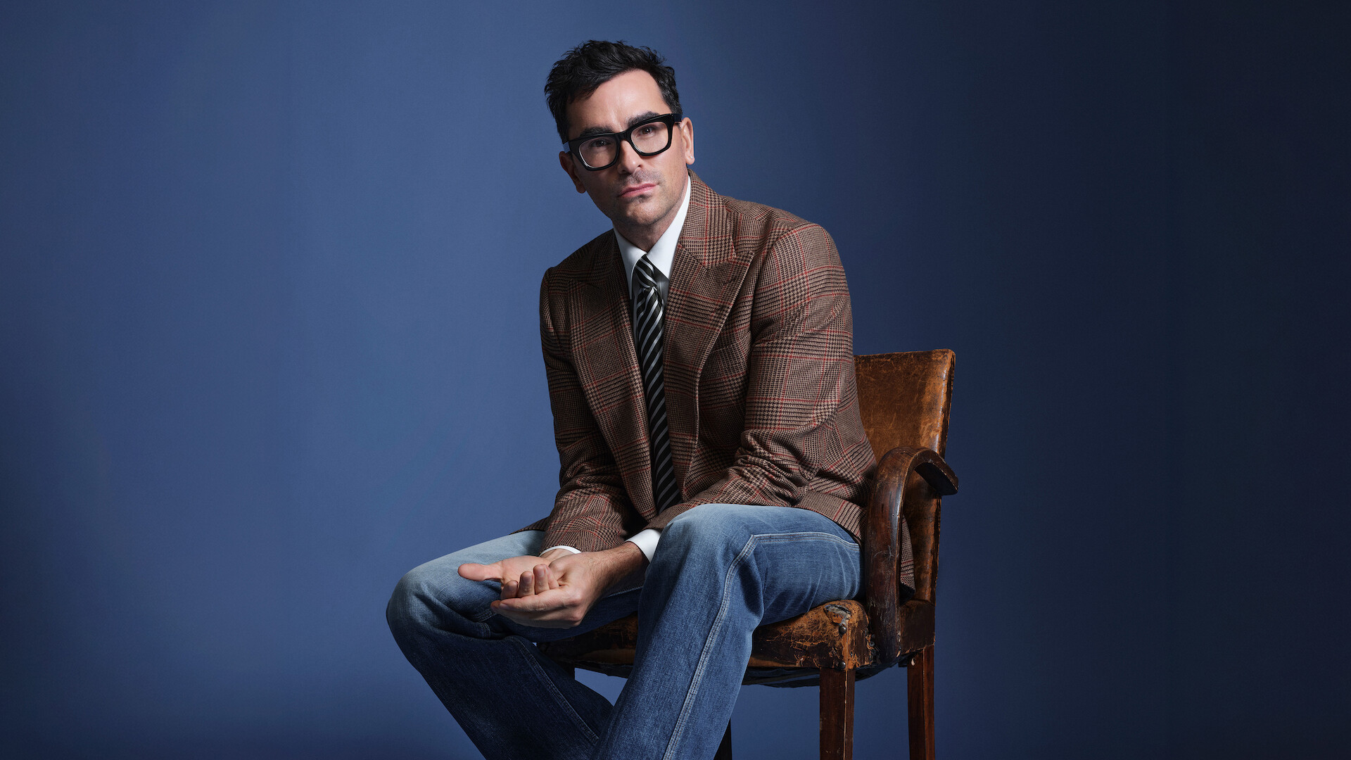 Dan Levy sits in a chair as he poses against a blue backdrop, wearing a checked blazer.