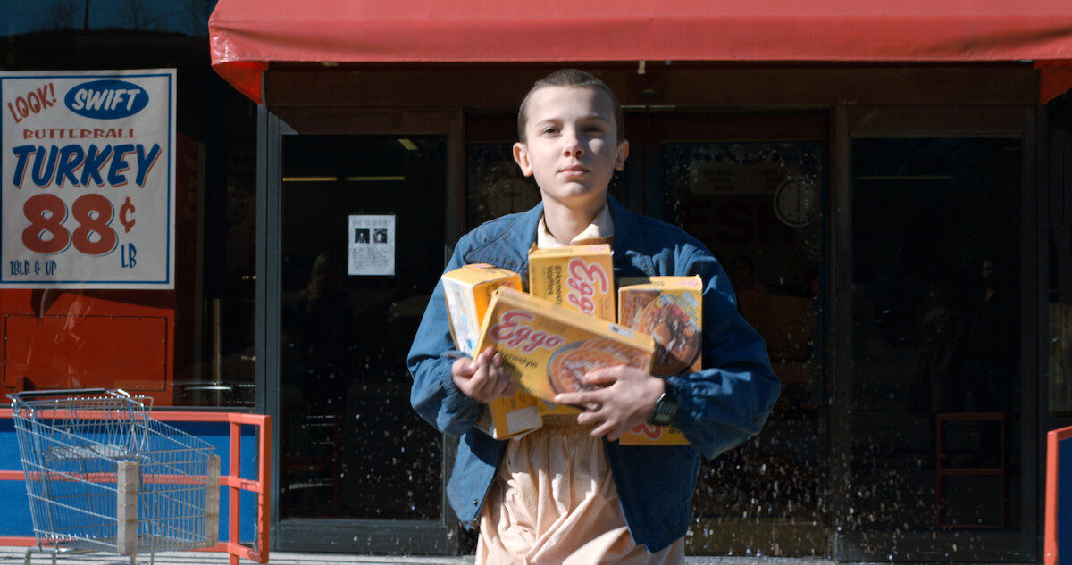 Eleven walks out of a grocery store holding several boxes of frozen waffles stacked in their arms, with a shopping cart and a vintage turkey-price sign visible in the background.