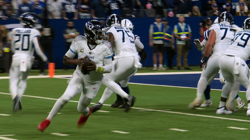 Football quarterback in white uniform running with the ball on a green field during a game, with teammates blocking and stadium spectators in the background.